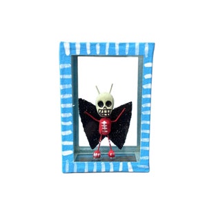 May include: A small, skeletal figure with red wings and a white skull is displayed in a blue and white frame. The figure is standing on a small, black base.