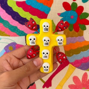 Puede incluir: Una cruz de madera amarilla con seis caras de calavera blancas y seis corazones rojos. La cruz está decorada con un diseño tradicional de arte popular mexicano.