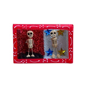 May include: A red wooden frame with a glass front, containing two small skeleton figures with painted faces. The background is red and white glitter, with blue and gold stars.