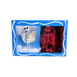 May include: A blue wooden frame with a white and red skeleton figure inside. The skeleton on the left is white with silver wings and the skeleton on the right is red with red wings. The background is a blue sky with white clouds.