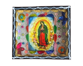 May include: A framed image of the Virgin Mary with a gold and silver glitter background. The frame is decorated with colorful flowers and stars.