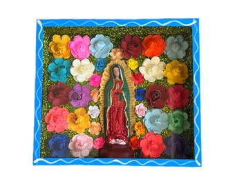 May include: A framed artwork featuring a colorful depiction of the Virgin Mary surrounded by paper flowers in various shades of pink, red, yellow, blue, and green. The frame is a bright blue with a wavy white border.