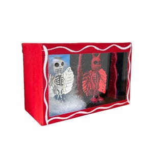 May include: A red box frame with a white border houses two skeleton figures. One is white with a silver wing, the other is red with horns. The background is blue and black, with a white, fluffy base.