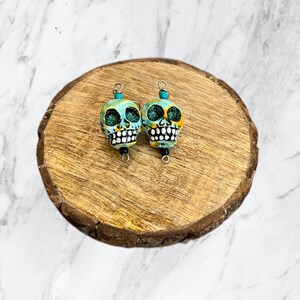May include: A pair of turquoise and yellow sugar skull earrings with black details. The earrings are on a wooden surface.