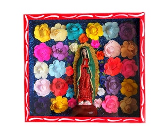 May include: A framed religious artwork featuring a statue of the Virgin Mary, surrounded by colorful paper flowers. The frame is red with a blue glitter background. The statue is in a green and red robe.