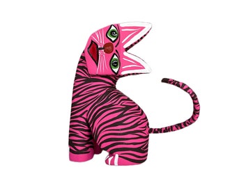 May include: A pink and black striped wooden cat figurine with a painted face. The cat has green eyes, a red nose, and a white mouth.