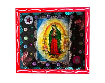May include: A framed image of the Virgin Mary with a red, white, and blue border. The image is surrounded by sequins and other decorative elements.