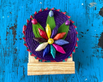 May include: A colorful, handmade, woven flower with green leaves, on a purple background, with a wooden stand. The flower is made of colorful, woven strips of material.