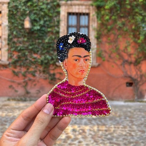 Op de afbeelding: Een roze, met pailletten bezet portret van Frida Kahlo met een zwart hoofdband en witte bloemen.