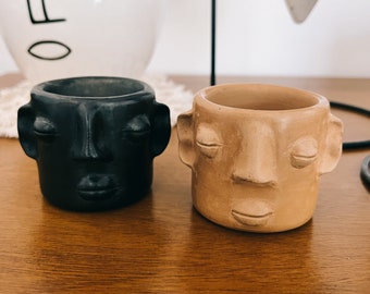 Puede incluir: Dos pequeñas macetas de cerámica con forma de cara. La maceta de la izquierda es negra con los ojos cerrados y una expresión neutral. La maceta de la derecha es marrón claro con los ojos cerrados y una expresión sonriente.