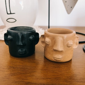 May include: Two small ceramic planters shaped like faces. The planter on the left is black with closed eyes and a neutral expression. The planter on the right is light brown with closed eyes and a smiling expression.