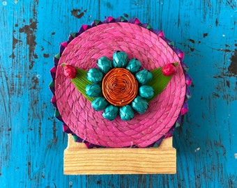 May include: A decorative, handcrafted item featuring a vibrant pink woven circular base. It is adorned with teal and orange woven accents, green leaves, and red berry-like details. The piece is mounted on a small, natural wood base.