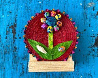 May include: A colorful flower made of woven straw with a red base and green leaves. The flower is mounted on a small wooden stand.