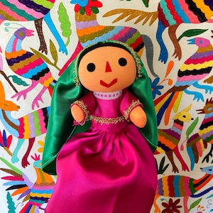 May include: A handmade doll wearing a pink dress and a green shawl. The doll is sitting in front of a colourful embroidered background with a variety of birds and flowers.
