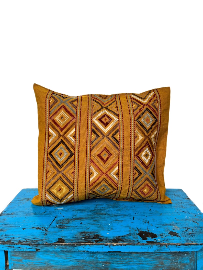 May include: A mustard yellow throw pillow with a geometric pattern in shades of brown, gray, and white. The pillow is sitting on a blue painted wooden surface.