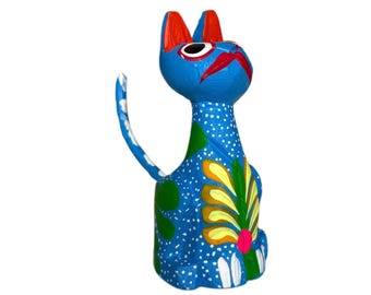 Könnte beinhalten: Eine lebendig bemalte Holzkatzenfigur. Die Katze ist überwiegend blau mit weißen Punkten, roten Ohren und einem roten Schnurrbart. Sie hat ein Blumendesign in Gelb, Grün und Pink. Der Schwanz ist gebogen, und die Katze sitzt.