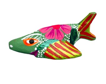 Könnte beinhalten: Handbemalte Holzfischfigur. Der Fisch ist hauptsächlich grün mit roten und gelben Streifen auf den Flossen und dem Schwanz. Auf dem Körper ist eine große rosa Blume gemalt, und weiße Punkte akzentuieren das Design.