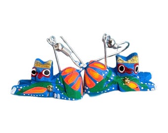 May include: A pair of handcrafted, blue wooden cat earrings. Each cat has a gold crown, red eyes, and orange and green accents. The earrings are connected by silver-colored metal wires.