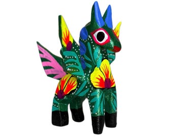 May include: A hand-painted, wooden alebrije figurine of a mythical creature. The figurine features a green body with yellow, red, and pink floral designs. It has blue horns, wings, and black feet. The eyes are large and white with a red outline.