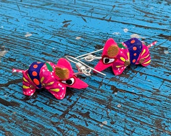 May include: A pair of handcrafted, vibrant pink armadillo earrings. Each earring features a detailed design with gold, blue, yellow, and red accents. The earrings have silver-colored ear wires and are set against a blue, weathered wooden surface.