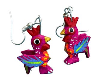 May include: A pair of handcrafted, magenta-colored rooster earrings. Each earring features a red comb, yellow beak, and blue and green wing accents. The earrings are made of wood and have silver-colored ear wires.