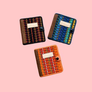 May include: Three small notebooks with different patterned covers. The covers are made of a woven fabric with a brown leather border. Each notebook has a blank label on the front and a black button closure.