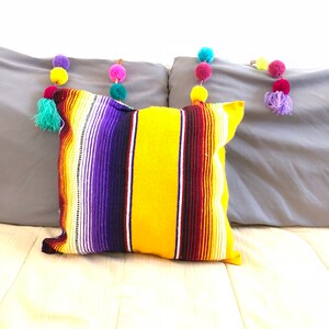 Könnte beinhalten: Ein gelbes, violettes und rotes gestreiftes Kissen mit Fransenrand. Das Kissen liegt auf einem grauen Sofa mit bunten Pompons, die über die Rückenlehne gespannt sind.