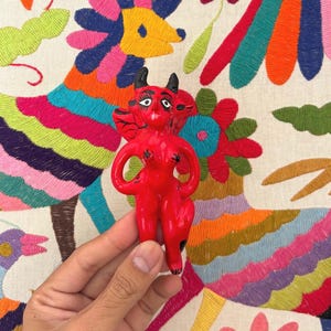 Puede incluir: Una figurita de cerámica roja de un diablo con cuernos y alas. La figurita está de pie sobre un fondo de tela bordada de colores.