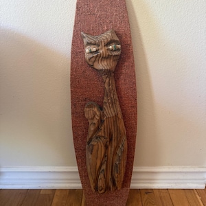 May include: A vertical wall plaque featuring a carved wooden cat figure mounted on a textured, rust-colored fabric backing. The cat has a natural wood grain finish, elongated features, and small green accents in the eyes, set against a tall, tapered board.