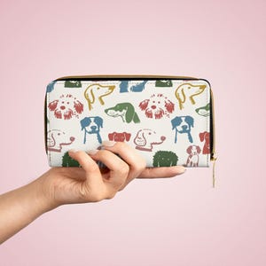 Könnte beinhalten: Eine weiße Geldbörse mit goldenem Reißverschluss und einem farbenfrohen Muster verschiedener Hunderassen. Die Hunde sind in Rot-, Grün-, Blau- und Gelbtönen gehalten. Die Geldbörse ist rechteckig und hat einen schwarzen Rand.