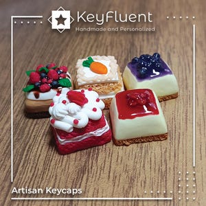 May include: Five miniature cake keycaps, each with a different design and frosting. The keycaps are made of polymer clay and are designed to look like real cakes. The keycaps are arranged in a row on a wooden surface. The keycaps are all different colors and have different toppings, including berries, whipped cream, and chocolate.