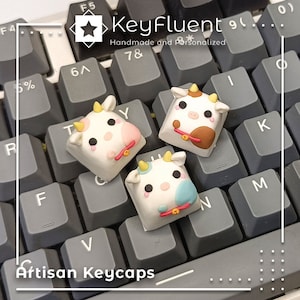 Könnte beinhalten: Drei weiße, kuhförmige Keycaps mit braunen Flecken, rosa und blauen Akzenten und gelben Hörnern. Die Keycaps befinden sich auf einer grauen Tastatur mit dem Text "KeyFluent Handmade and Personalized" und "Artisan Keycaps".