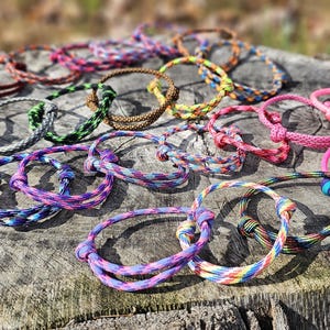 May include: A collection of numerous cord bracelets in a wide variety of solid colors and multi-colored braided patterns. Each bracelet features a simple adjustable sliding knot closure mechanism, allowing for flexible sizing and easy wearability.