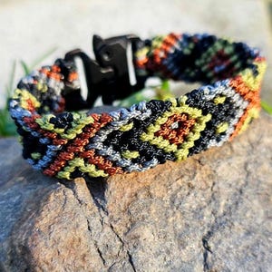 May include: A woven bracelet with a black clasp. The bracelet features a geometric pattern in shades of black, orange, yellow, gray, and blue. The bracelet is resting on a rock, showcasing its intricate design and color palette.