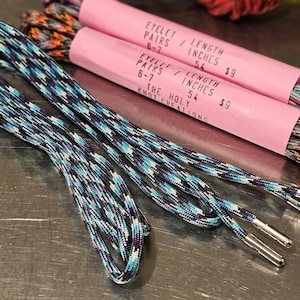 May include: Shoelaces with a blue, white, and purple pattern are displayed next to two pink paper rolls. The rolls have text that reads "EYELET PAIRS 6-7 / LENGTH INCHES 54 $9" and "THE HOLY KNOT CREATIONS."