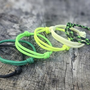 May include: Five colorful paracord bracelets in green, yellow, and black. The bracelets are tied in a knot and are lying on a wooden surface.