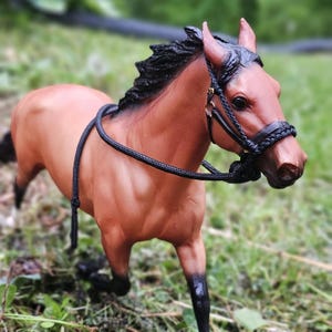 May include: A detailed figurine of a horse with a brown coat and black mane and tail. The horse is wearing a black bridle and rope halter. The horse is posed in a grassy area.