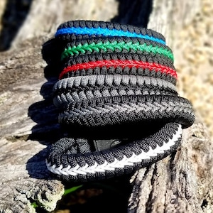 May include: A stack of five paracord bracelets in various colors, including black, gray, green, red, and blue. The bracelets are woven with a complex pattern and are stacked on top of each other.