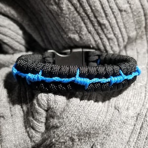 May include: Black and blue paracord bracelet with a black plastic clasp. The bracelet is made of tightly woven black cord with a bright blue accent woven through the center. The bracelet is resting on a gray, textured surface.
