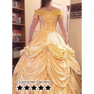 May include: A yellow ball gown with a lace bodice and a corset back. The skirt is full and has a bustle in the back. The gown is worn by a person standing in front of a bookshelf.