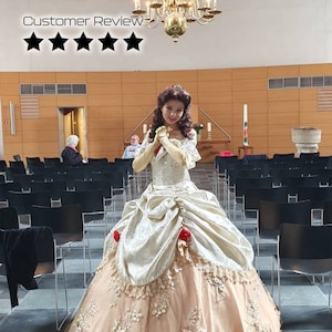 May include: A woman dressed as Belle from the Disney movie Beauty and the Beast. She is wearing a gold and white ball gown with a lace trim and red roses. She is standing in a church with rows of black chairs.