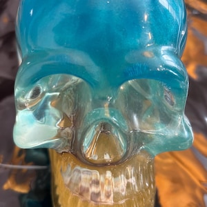 May include: A decorative skull sculpture with a gradient color scheme. The top of the skull is a vibrant teal, transitioning to a translucent, golden hue at the jawline. The skull's details are clearly defined, showcasing its unique design.