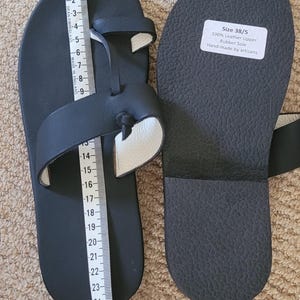 May include: A pair of black leather sandals with a toe loop and a rubber sole. A white measuring tape is placed along the length of one sandal, showing a measurement of approximately 25 centimeters. A label on the sole reads Size 38/5.