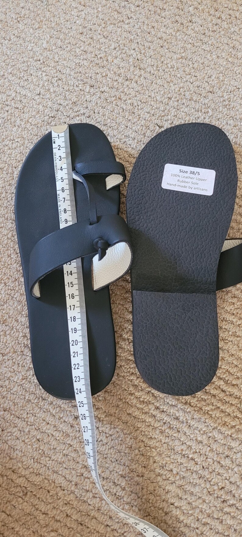 May include: A pair of black leather sandals with a toe loop and a rubber sole. A white measuring tape is placed along the length of one sandal, showing a measurement of approximately 25 centimeters. A label on the sole reads Size 38/5.