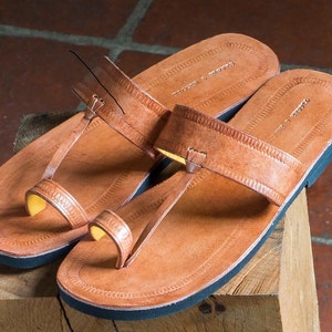 May include: A pair of tan leather sandals featuring a toe-post design and a wide horizontal strap across the top. The footbed is embossed with a decorative border pattern and the text "TRADERS & DROVERS". The sandals have a solid black sole.