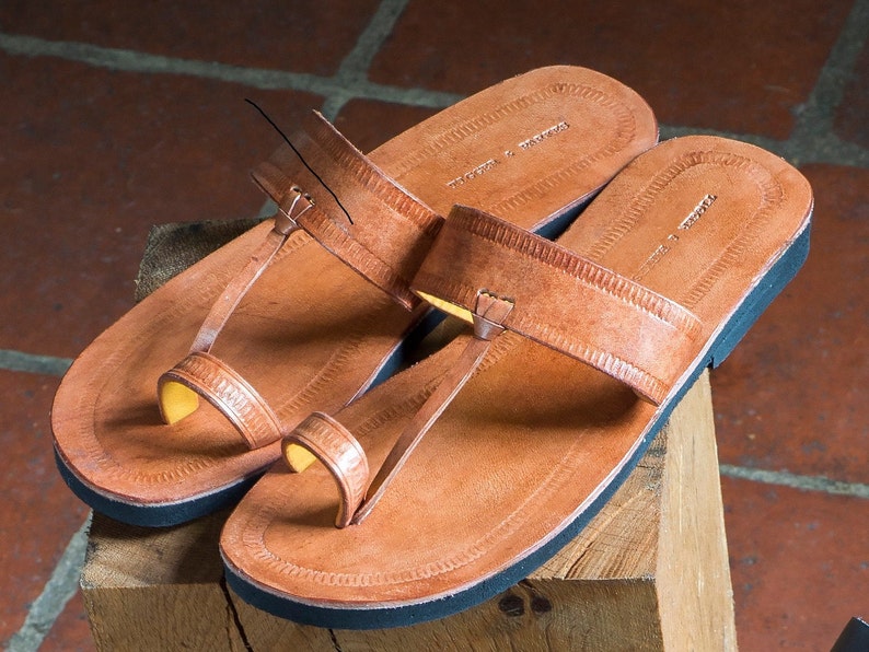 May include: A pair of tan leather sandals featuring a toe-post design and a wide horizontal strap across the top. The footbed is embossed with a decorative border pattern and the text "TRADERS & DROVERS". The sandals have a solid black sole.