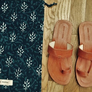 May include: A pair of tan leather toe-ring sandals with a flat sole and a matching dark blue fabric drawstring bag featuring a white botanical print and a white label with the text Tigger & Parkes printed in black on the lower left corner.