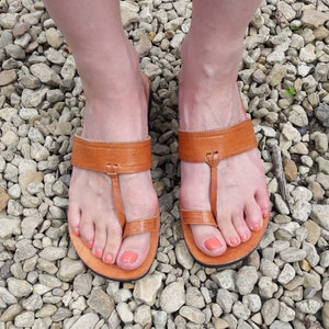 May include: A pair of tan leather sandals featuring a wide horizontal strap across the midfoot and a separate loop that secures the big toe. The sandals have a flat sole and a simple design with visible stitching along the edges of the leather straps.