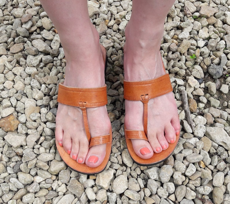 May include: A pair of tan leather sandals featuring a wide horizontal strap across the midfoot and a separate loop that secures the big toe. The sandals have a flat sole and a simple design with visible stitching along the edges of the leather straps.