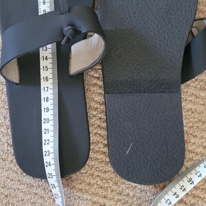 May include: A pair of black sandals with a toe loop and a knotted strap design. One sandal shows the number 39 written on the footbed. A white measuring tape is placed over the sandals, indicating a length of approximately 25 centimeters.